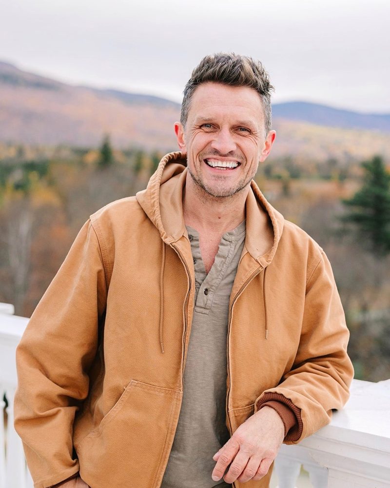 Alex Bond smiling on a balcony with autumn mountains behind him